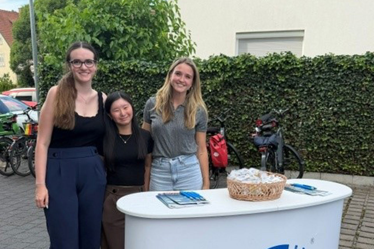 Drei junge Frauen stehen hinter einem Werbestand für „Gewinnsparen“ auf einem gepflasterten Außenbereich.