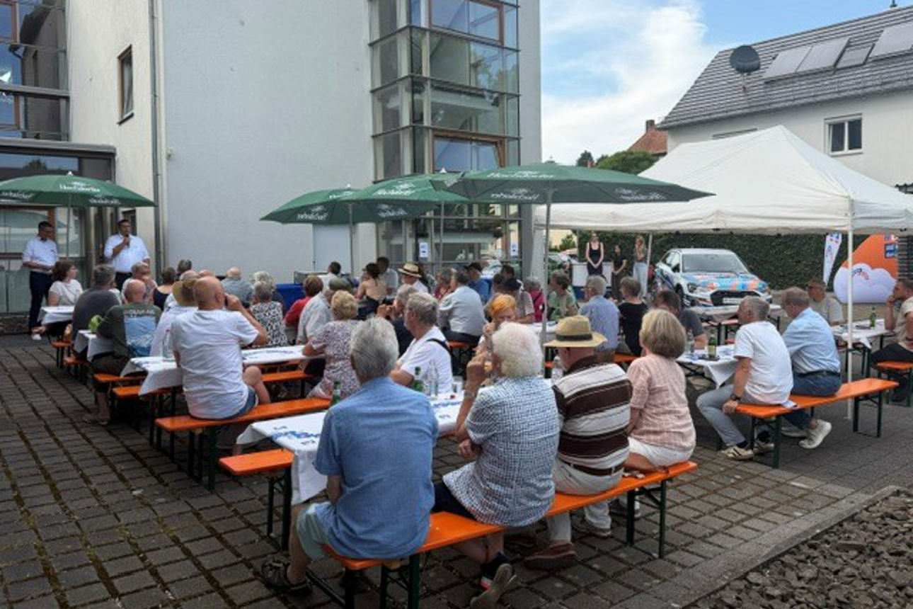 Eine große Gruppe von Menschen versammelte sich bei einer Veranstaltung im Freien, bei der Tische, Bänke und Sonnenschirme au
