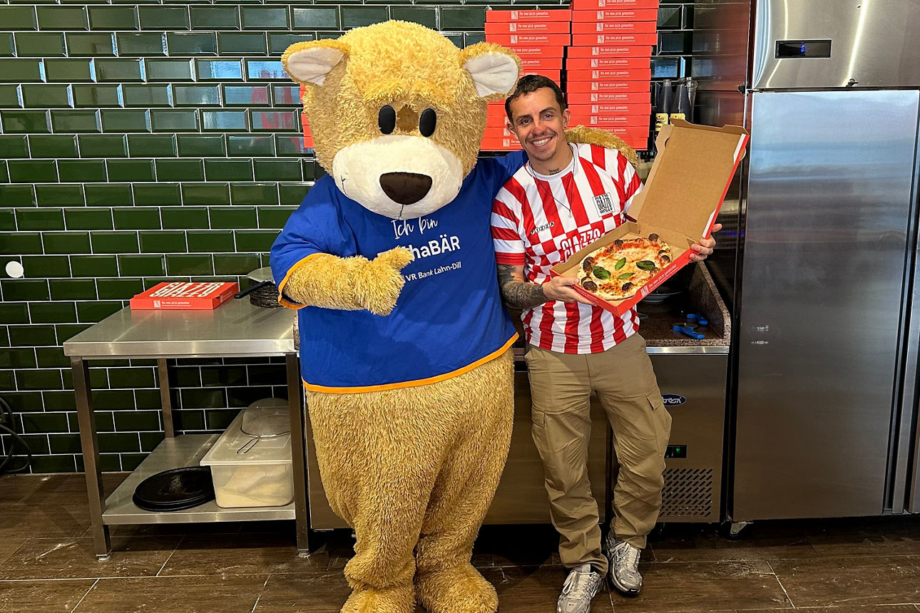 Großer Teddybär in blauem T-Shirt steht neben Person in rot-weiß gestreiftem Trikot, die eine offene Pizzaschachtel hält.