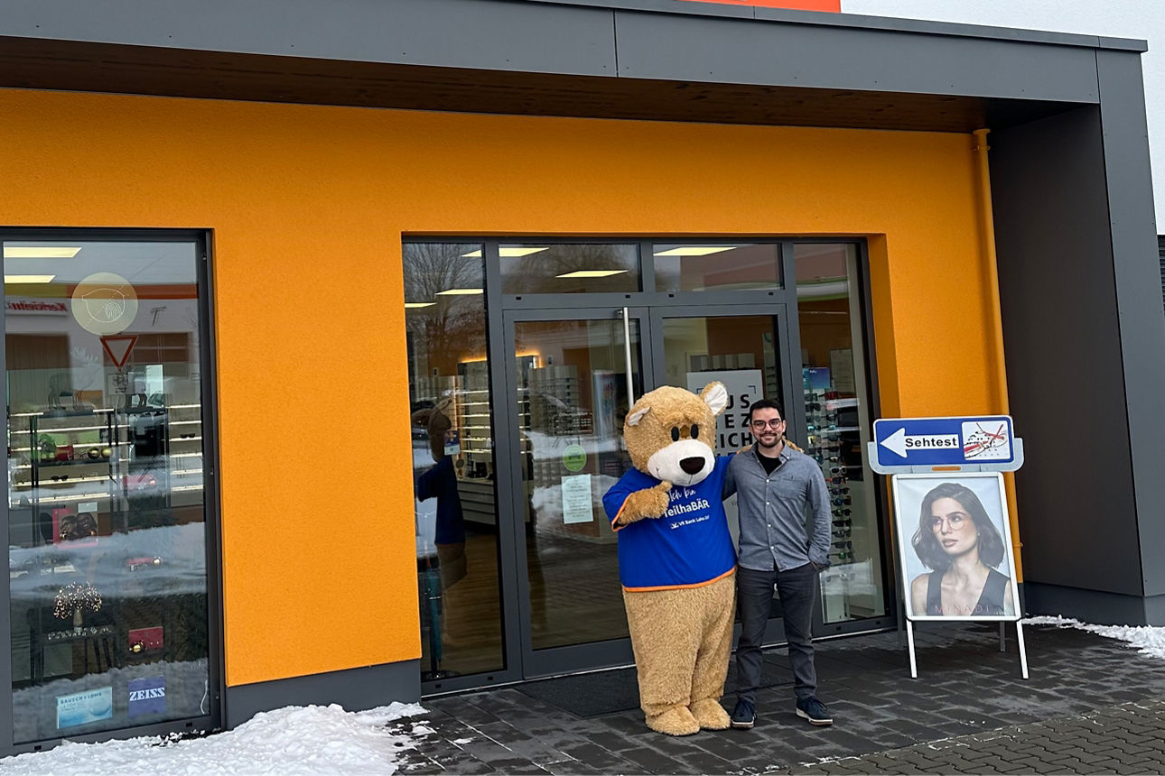 Vor einem orangefarbenen Geschäft mit der Aufschrift 'Optik LANG Gut Sehen' steht eine Person neben einer Bärenfigur in blauem T-Shirt.