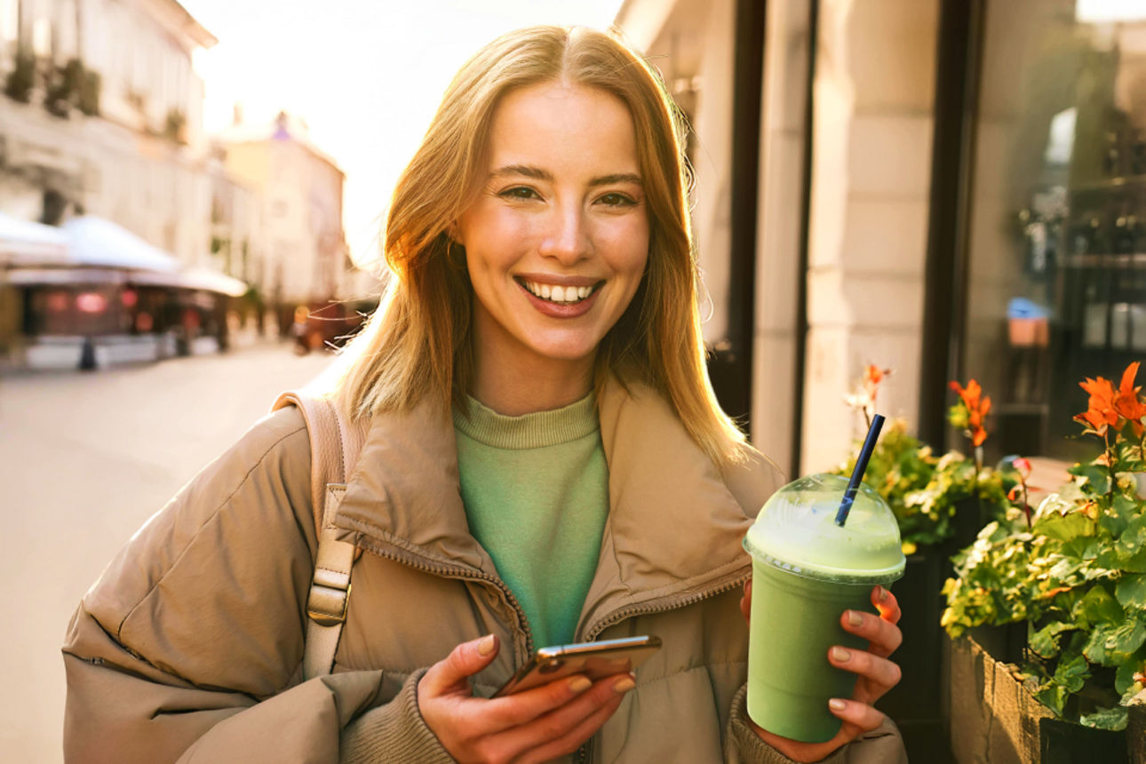 Frau lächelt mit Smartphone und Matcha in der Hand in die Kamera
