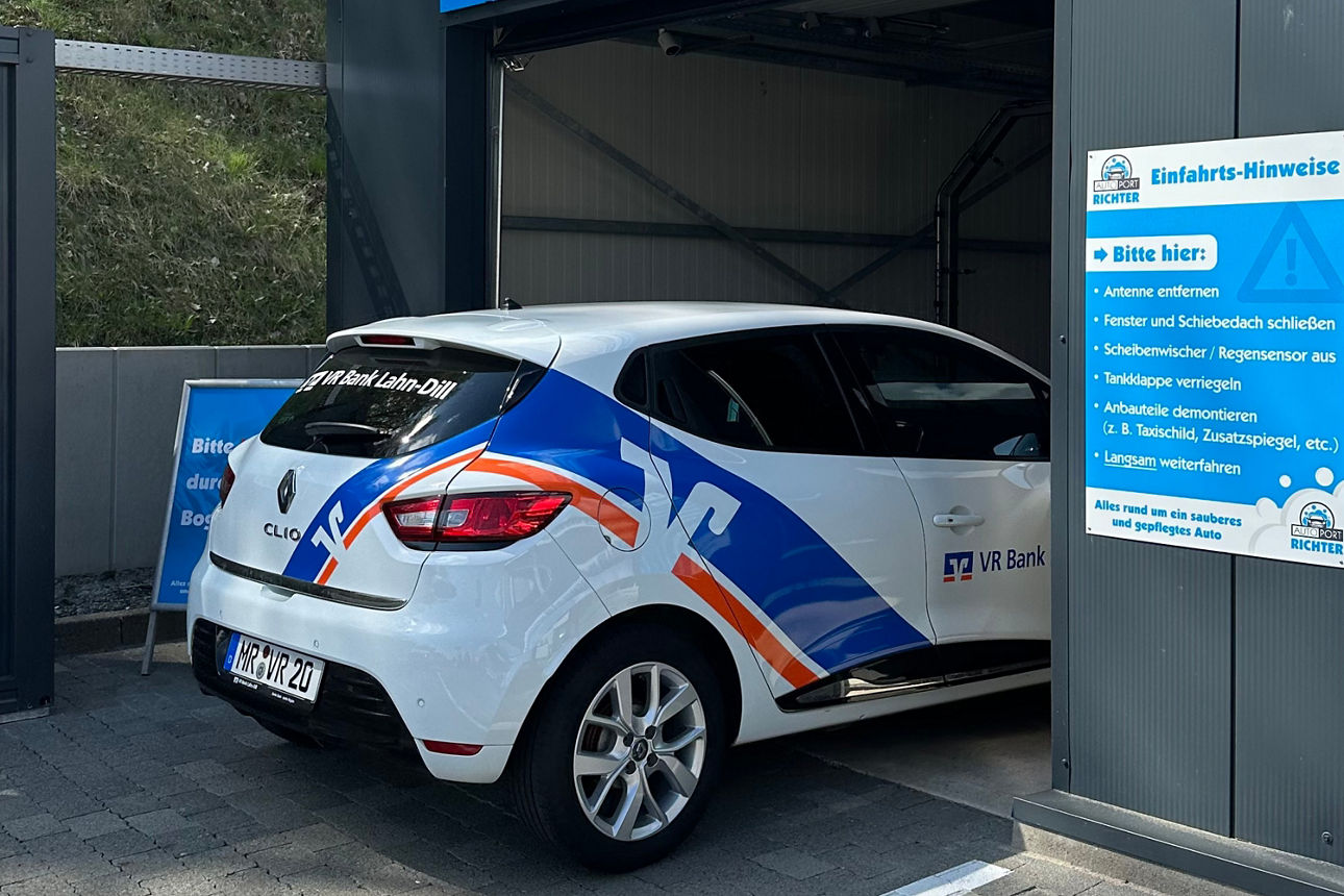 Weißer Renault Clio mit blau-orangen Werbeaufklebern steht in einer Waschanlage neben einem Schild mit Einfahrts-Hinweisen.