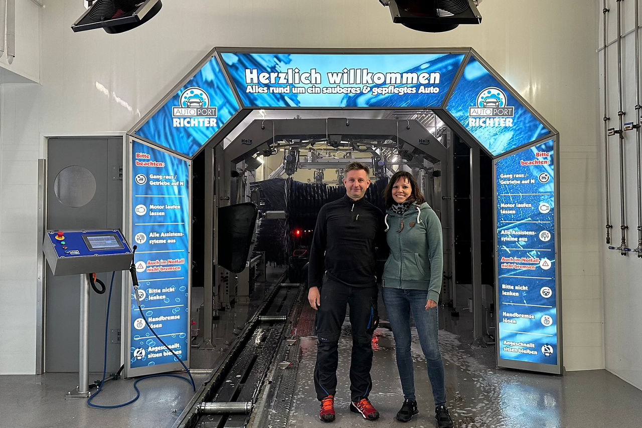 Zwei Personen stehen vor einem blauen Durchgang mit der Aufschrift 'Herzlich willkommen Alles rund um ein sauberes & gepflegtes Auto' in einer Autowaschanlage.