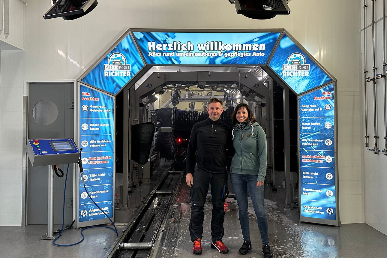 Zwei Personen stehen vor einem blauen Durchgang mit der Aufschrift 'Herzlich willkommen Alles rund um ein sauberes & gepflegtes Auto' in einer Autowaschanlage.