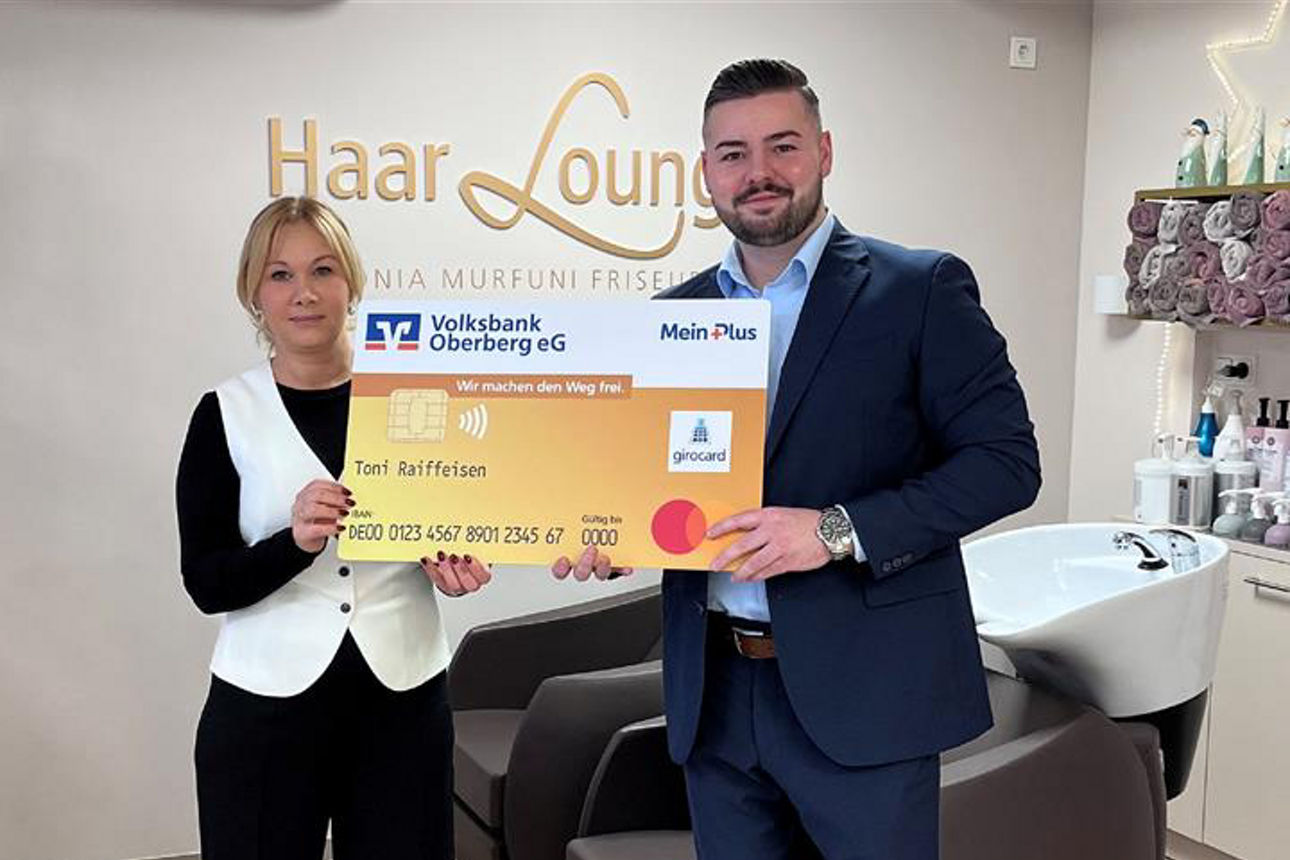 Ein Mann und eine Frau lächeln in die Kamera und halten gemeinsam eine übergroße, symbolische Bankkarte der Volksbank Oberberg.