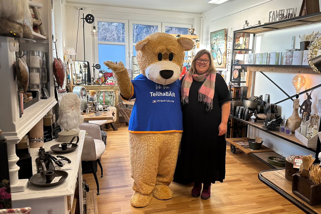 Person mit schwarzem Kleid und rosa Schal steht neben großem Teddybär-Maskottchen mit blauem T-Shirt in einem hellen Laden mit Holzfußboden und Regalen voller Dekorationsartikel