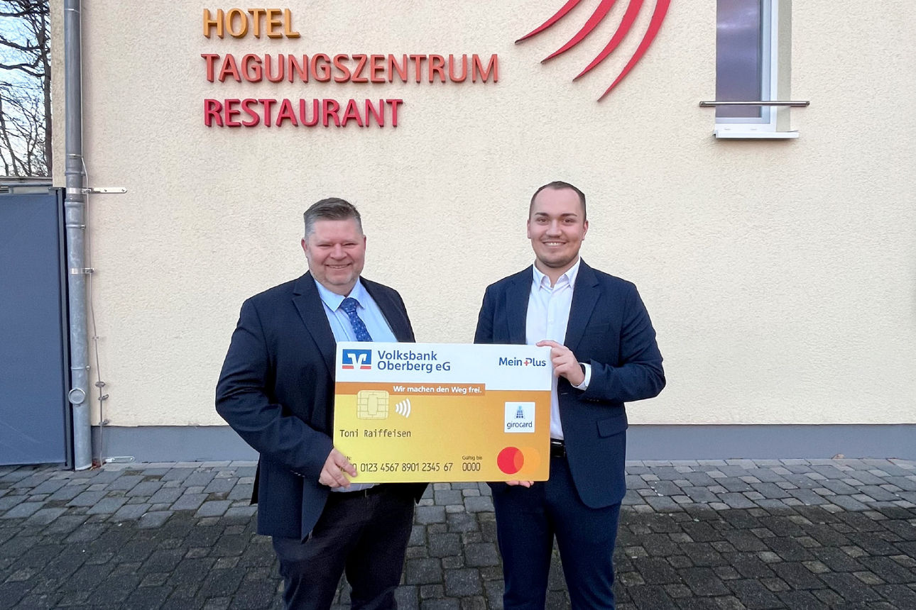 Zwei Männer in Anzügen stehen vor einem Gebäude mit der Beschriftung „Hotel Tagungszentrum Restaurant“. Sie lächeln in die Kamera und halten gemeinsam eine übergroße, symbolische Bankkarte der Volksbank Oberberg.