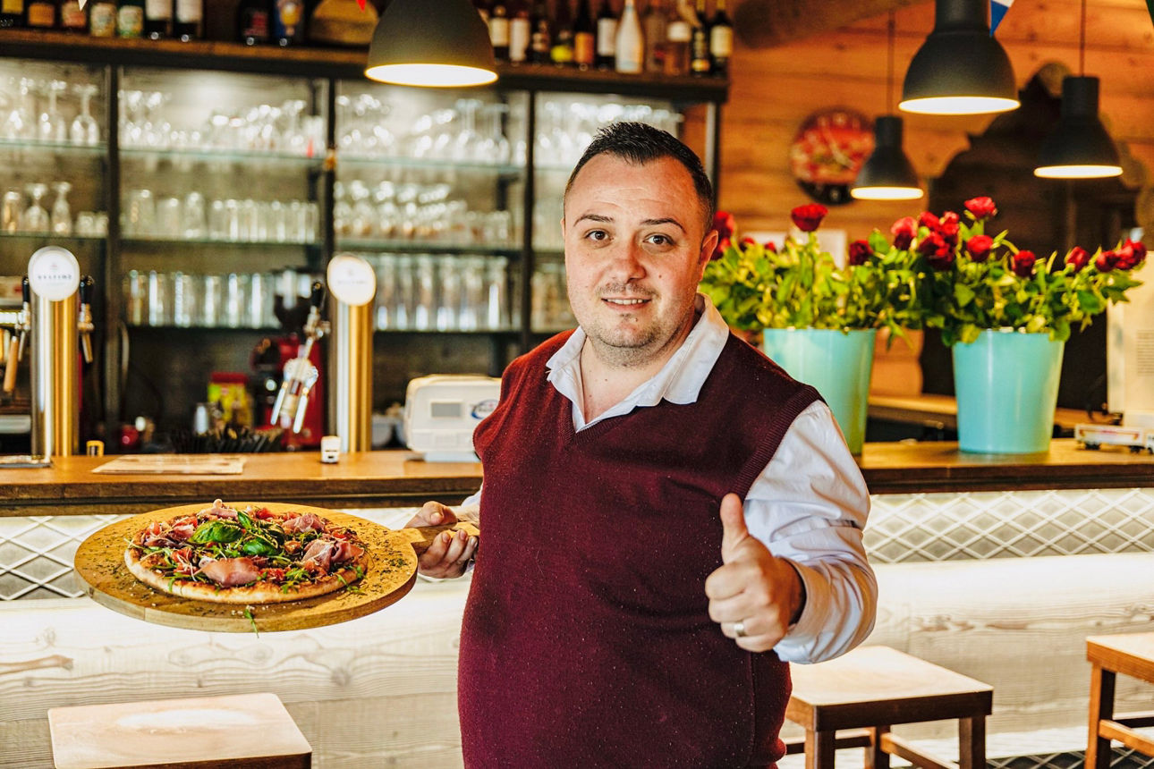 Mann in weißem Hemd und rotem Pullover hält eine Pizza auf einem Holzbrett und zeigt Daumen hoch in einem Restaurant mit Gläserregal und blauen Blumentöpfen im Hintergrund