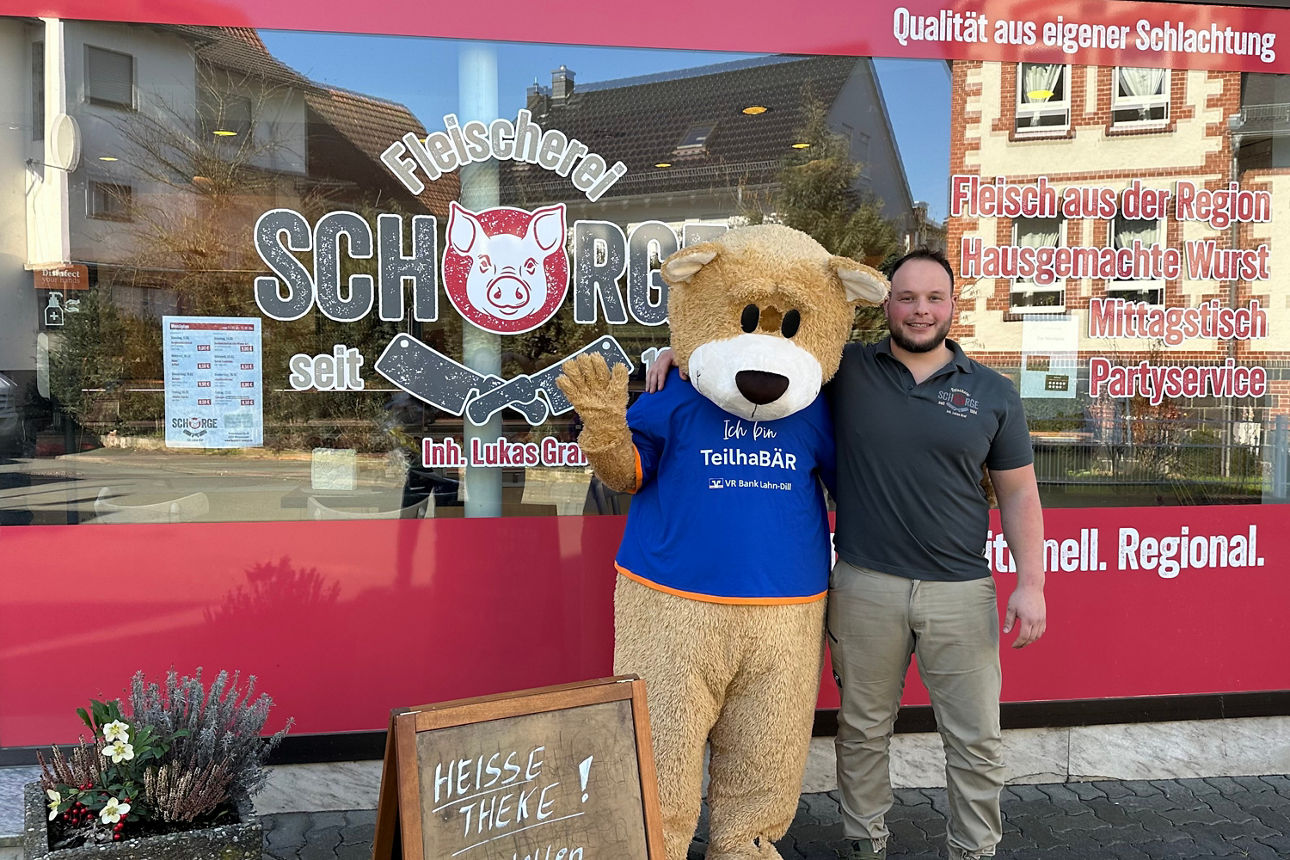 Ein Mann steht neben einer Person im Bärenkostüm vor der Schaufensterscheibe einer Fleischerei mit Werbetexten und einem Aufsteller mit der Aufschrift 'HEISSE THEKE'