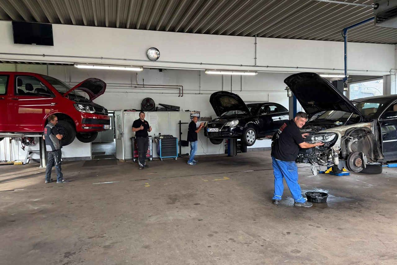 Vier Männer in einer Werkstatt an verschiedenen Autos auf Hebebühnen bei der Reparatur.