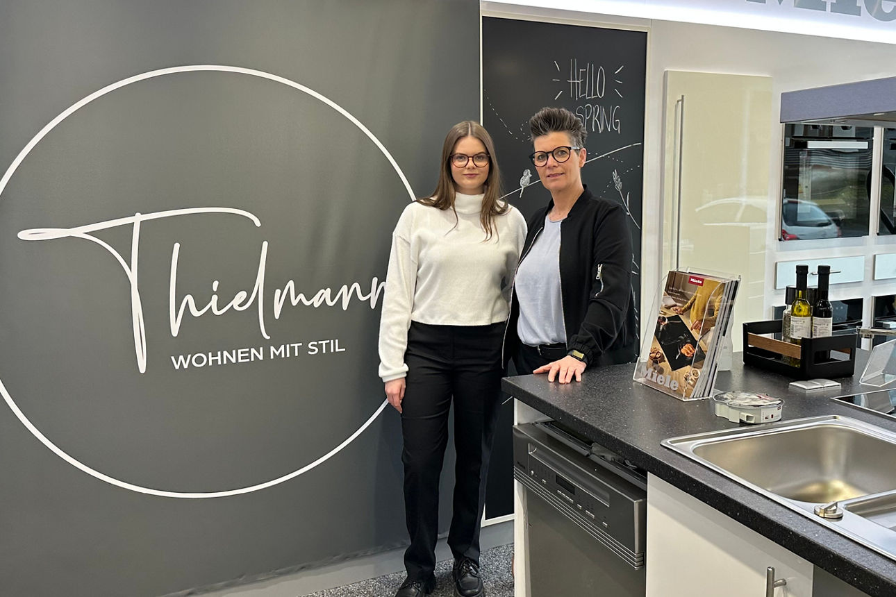 Zwei Personen stehen nebeneinander vor einer Wand mit dem Schriftzug 'Thielmann WOHNEN MIT STIL' in einer modernen Küchenausstellung.