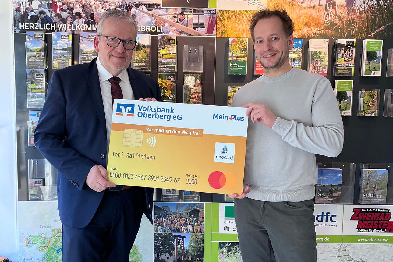 Zwei Männer lächeln in die Kamera und halten gemeinsam eine übergroße, symbolische Bankkarte der Volksbank Oberberg.