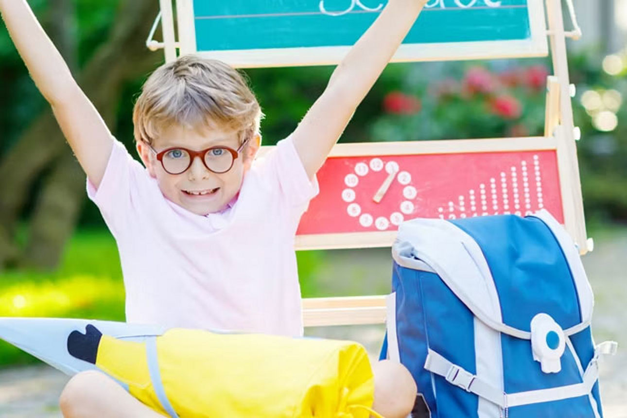 Ein glücklicher Junge mit Brille reißt die Arme hoch, umgeben von einer bunten Schultüte und einem blauen Schulranzen.