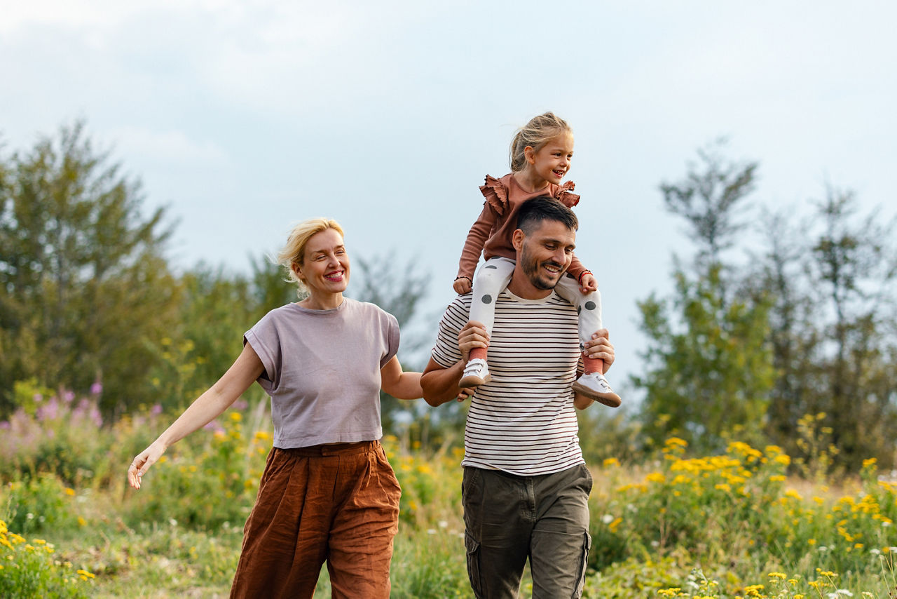 Familie geht in der Natur spazieren, der Vater trägt die kleine Tochter auf den Schultern, alle lachen