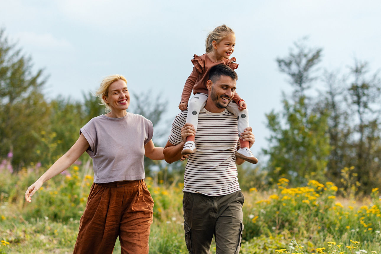 Familie geht in der Natur spazieren, der Vater trägt die kleine Tochter auf den Schultern, alle lachen