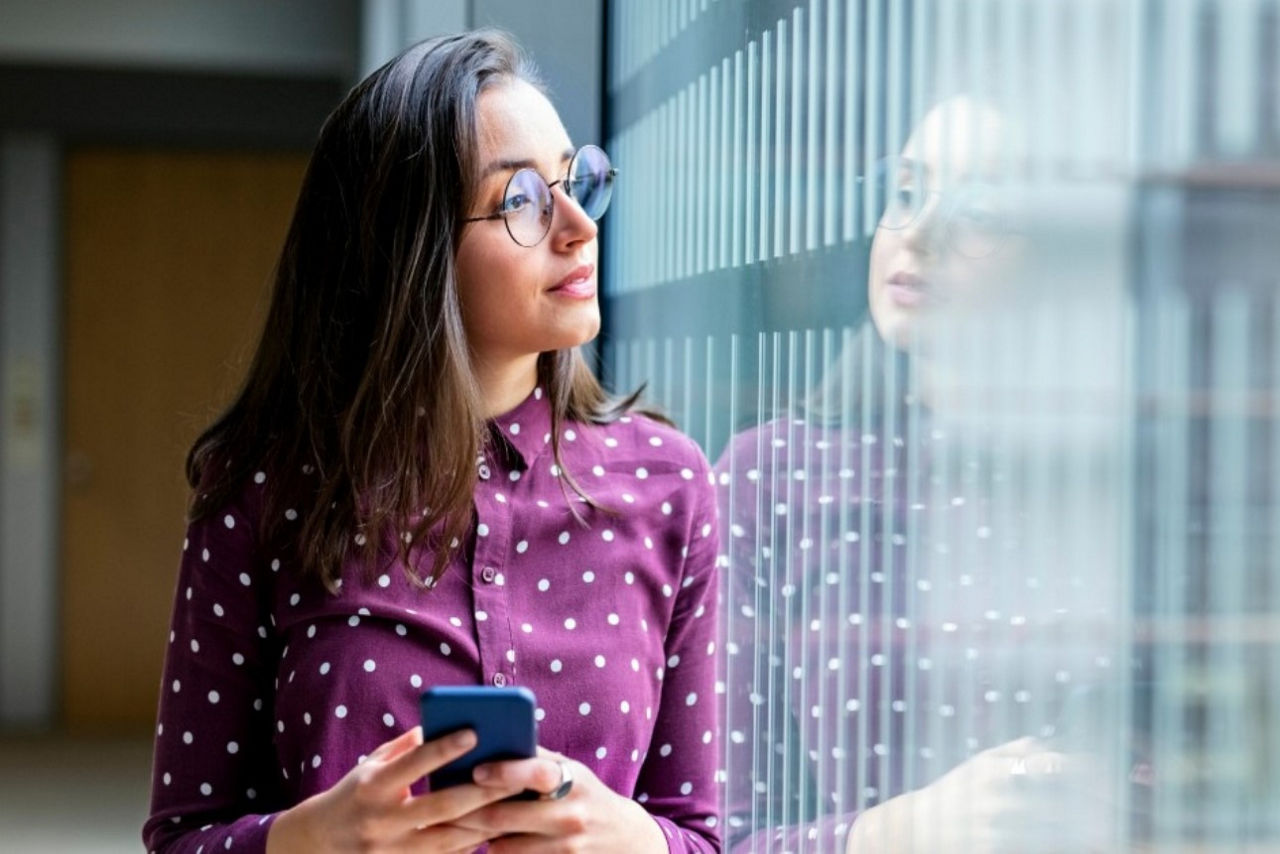 Frau, die ihr Vermögen über der BankingApp stets im Blick hat, blicktaus dem Fenster entspannt ihrer Zukunft entgegen