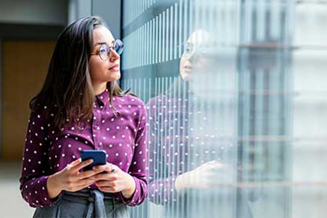 Frau steht gelehnt an ein Fenster, schaut raus mit Handy in der Hand