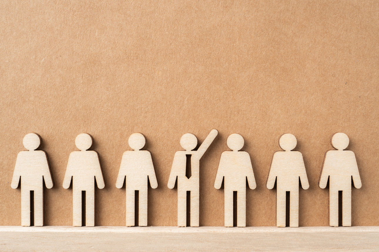 Sieben stilisierte Holzfiguren in einer Reihe, wobei eine Figur mit einer Krawatte hervorsticht und den Arm hebt.