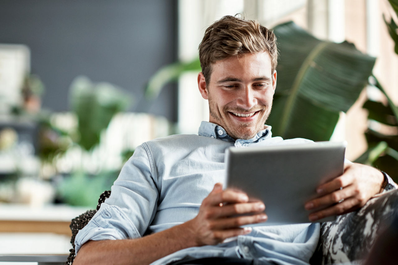 Auf dem Bild sitzt ein Mann auf der Couch und blickt auf ein Tablet in seiner Hand.