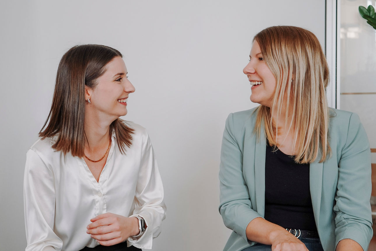 Zwei Bankmitarbeiterinnen sitzen nebeneinander und lächeln sich an 