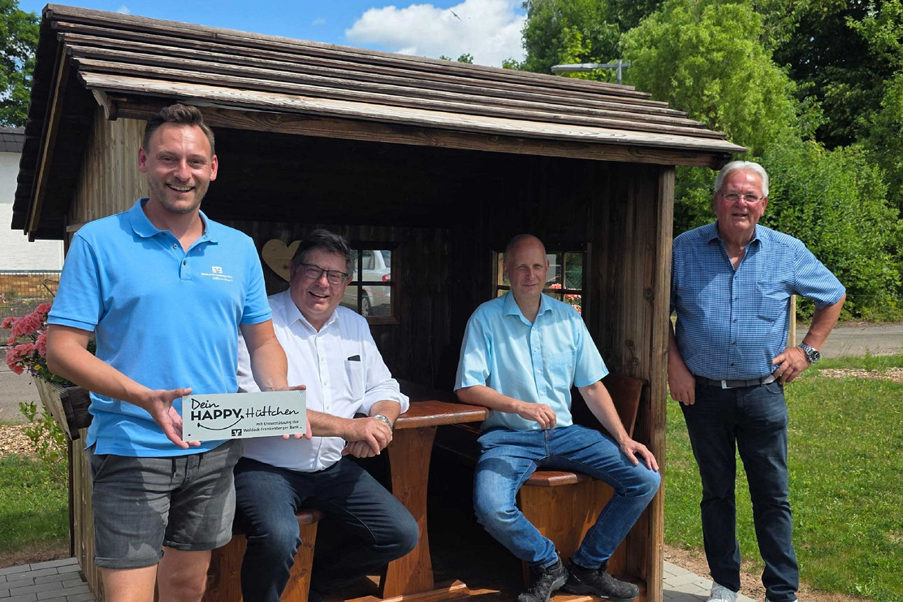Vier Männer lächeln an und in einer hölzernen Rast-Schutzhütte; einer hält ein Schild mit der Aufschrift „Dein HAPPY, Plätzch