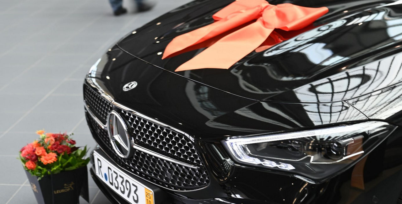 Auf dem Bild ist ein dunkles Auto mit einer Schleife zu sehen. Vor dem Auto stehen bunte Blumen.