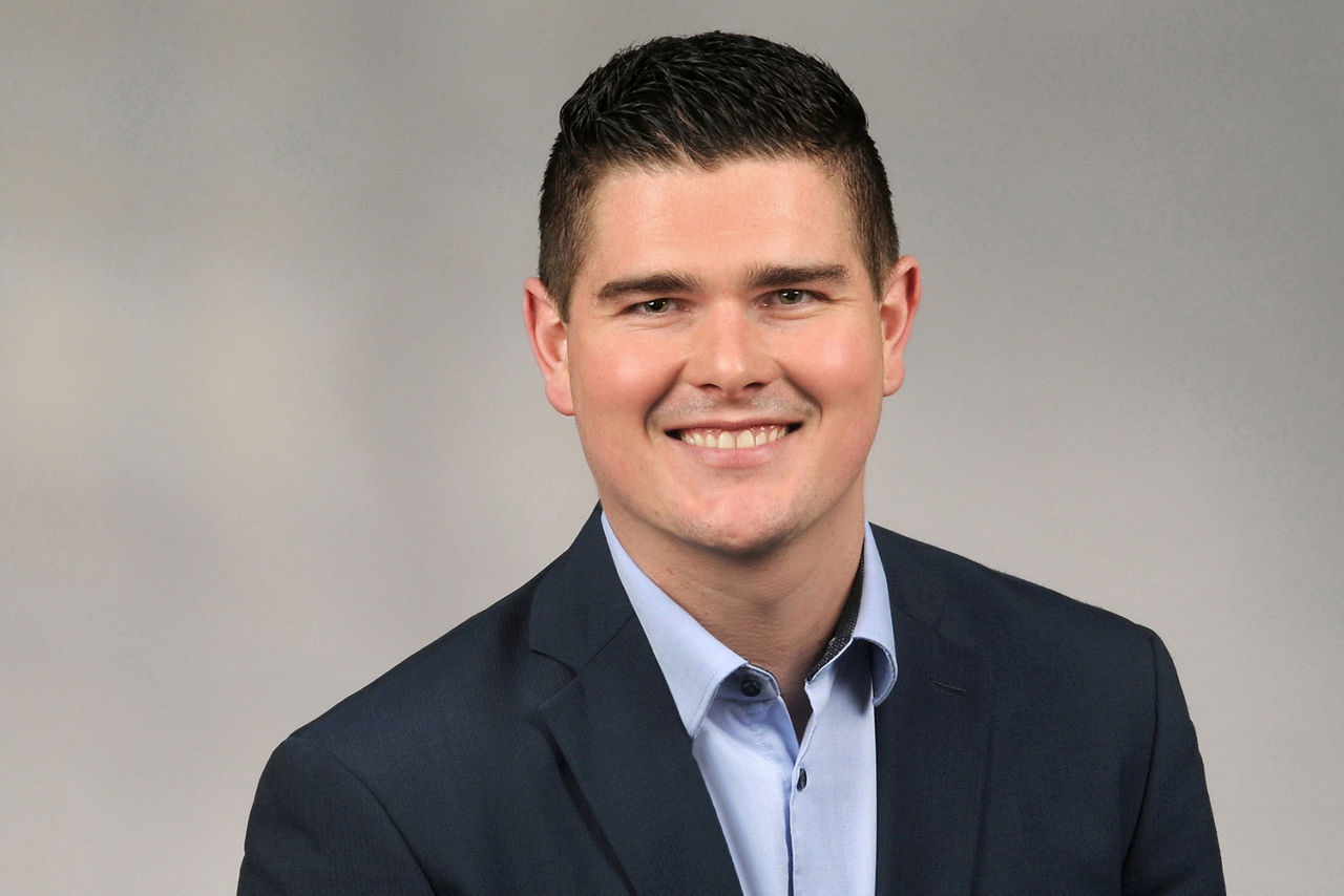 Mann mit dunklem kurzen Haar trägt dunkelblauen Anzug und hellblaues Hemd vor neutralem Hintergrund