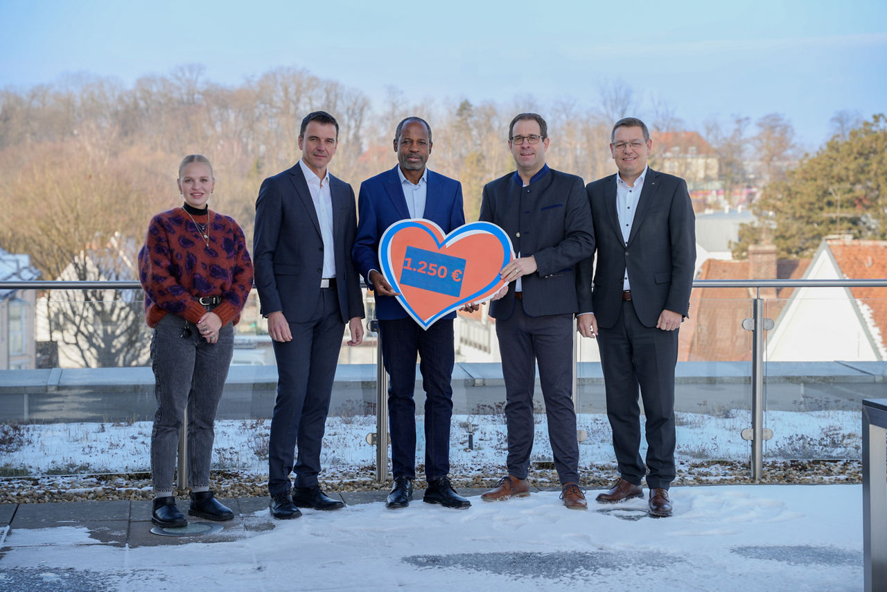 1 junge Frau und 4 Männer stehen auf verschneiter Terrasse entlang eines Geländers. 2 Herren halten ein orangenes Herz mit der Aufschrift 1.250 Euro. Im Hintergrund Hausdächer und Bäume