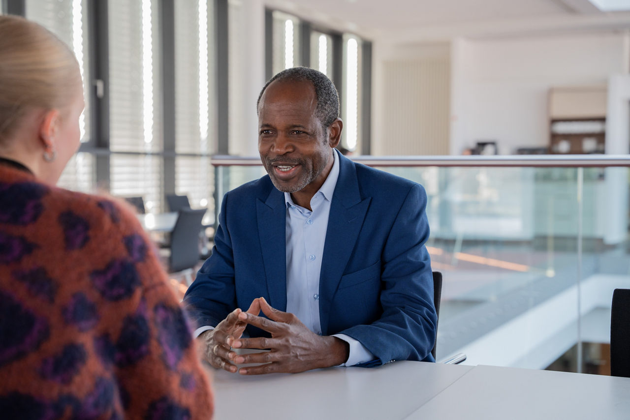 Mann im blauen Sakko und hellem Hemd sitzt im Gespräch an einem Tisch in modernem Gebäude ; gegenüber im Anschnitt von hinten zu sehen eine junge blonde Frau