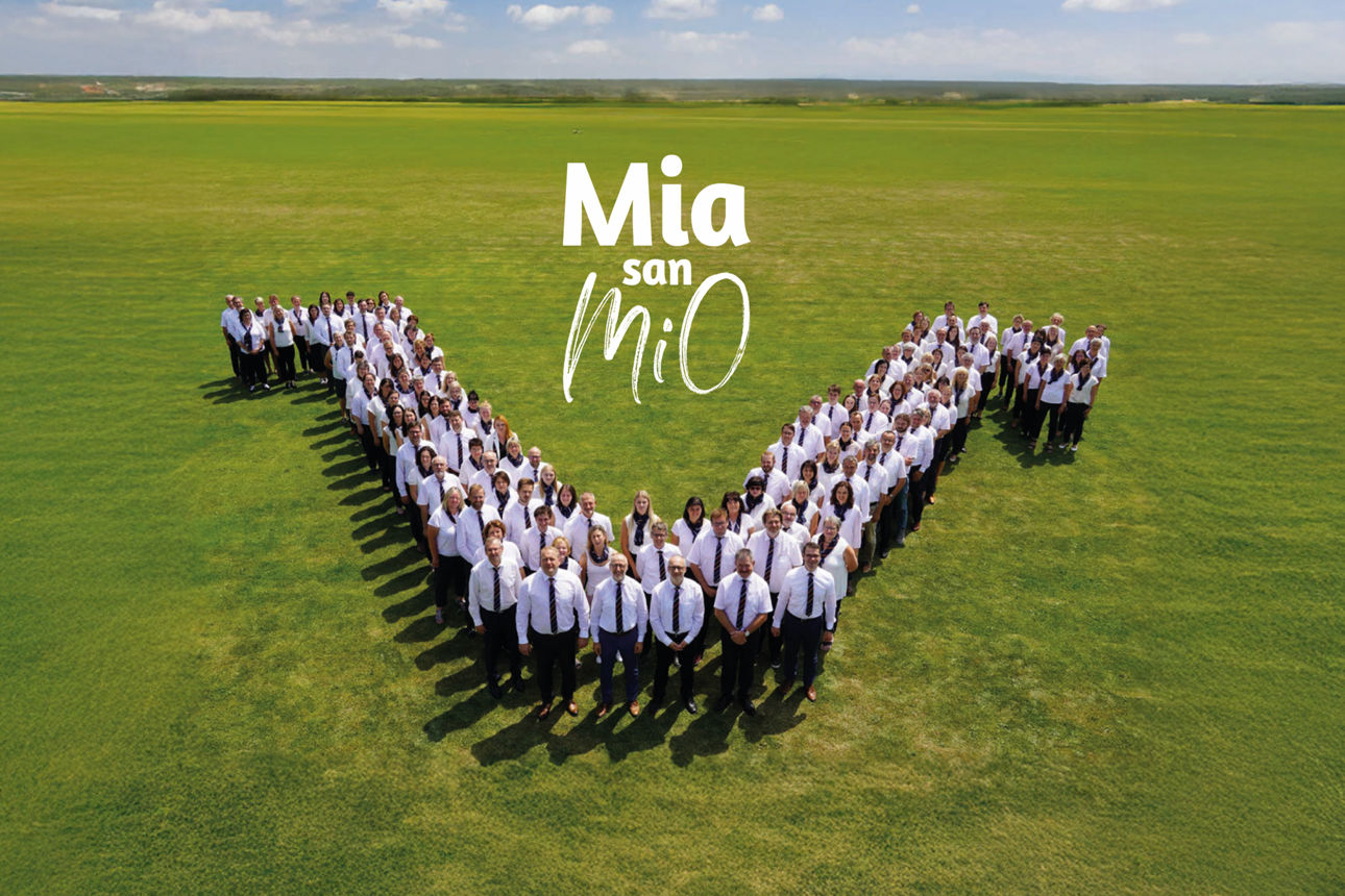Viele Mitarbeiter bilden ein V auf einer Wiese mit Text Mia san MiO