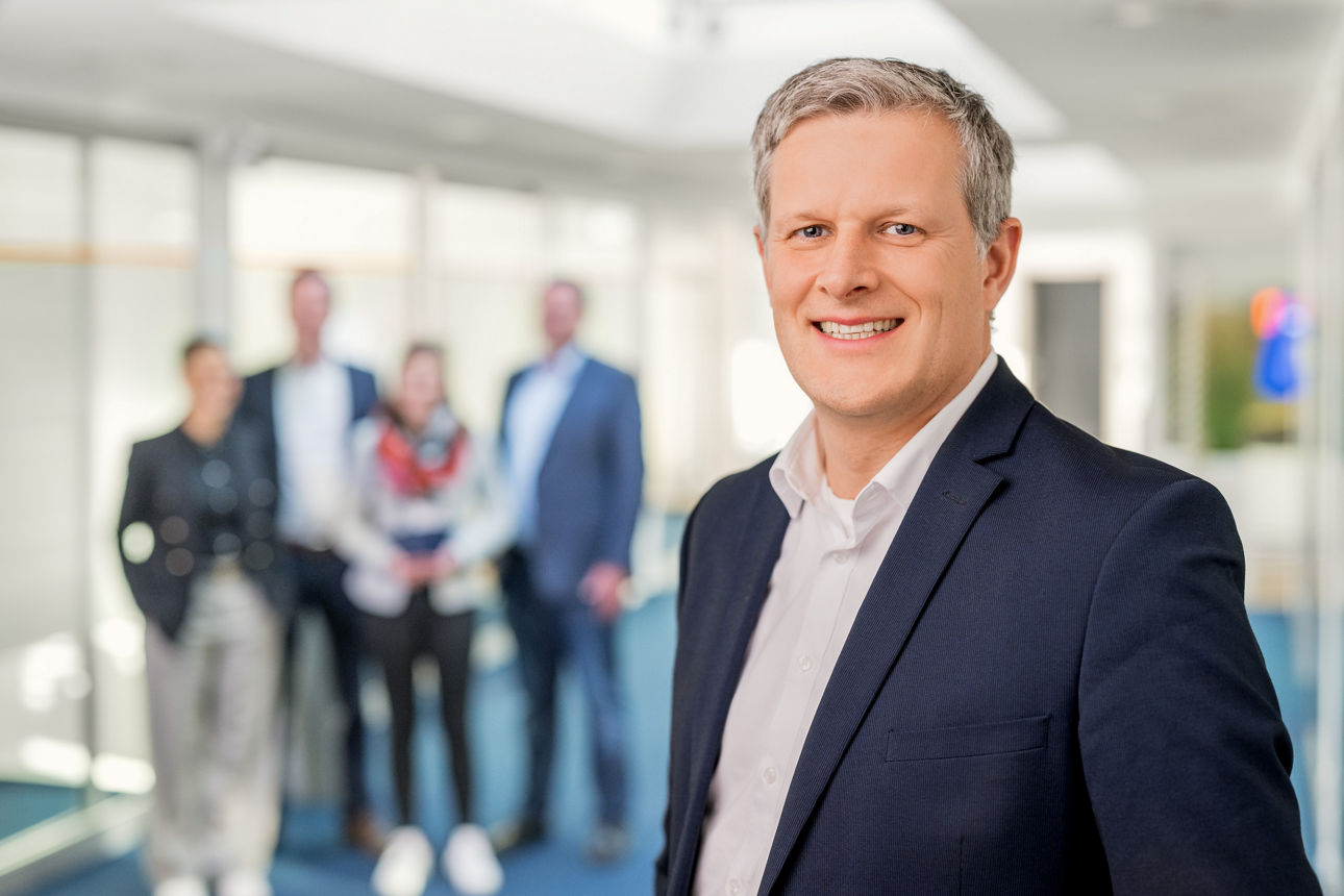 Ein Mann steht lächelnd und das Team hinter ihm.