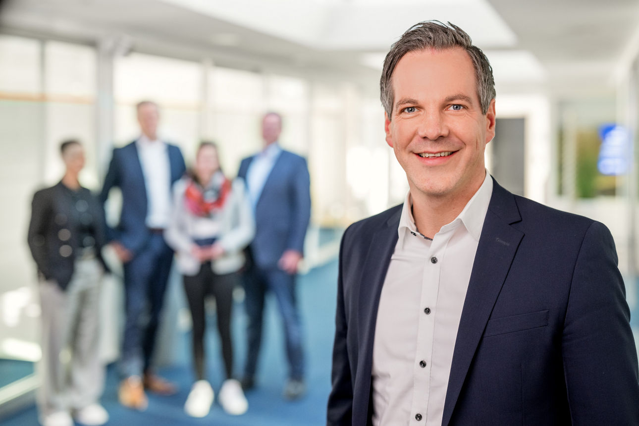 Michael Zobel lächelt in die Kamera und im Hintergrund steht das Team der Firmenkundenabteilung