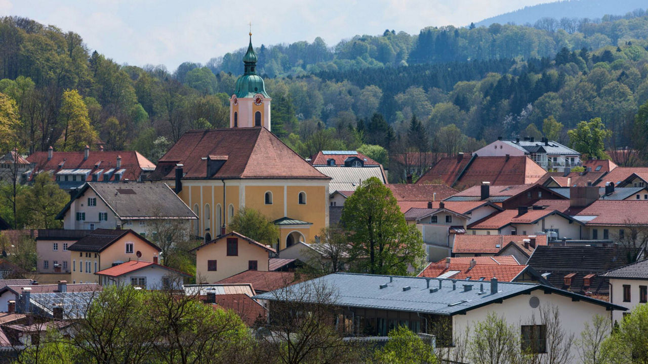 Eine malerische Ansicht der Stadt Miesbach mit einer großen, gelben Kirche mit grünem Zwiebelturm in der Mitte. Sie ist umgeben von zahlreichen Häusern mit roten Dächern, eingebettet in eine grüne, bewaldete Hügellandschaft.