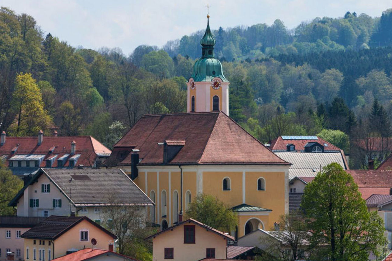 Eine malerische Ansicht der Stadt Miesbach mit einer großen, gelben Kirche mit grünem Zwiebelturm in der Mitte. Sie ist umgeben von zahlreichen Häusern mit roten Dächern, eingebettet in eine grüne, bewaldete Hügellandschaft.
