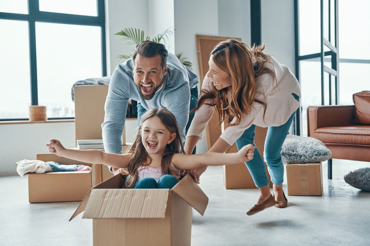 Eltern freuen sich zusammen mit Kind in einem Karton sitzend über den Bezug der neuen Wohnung