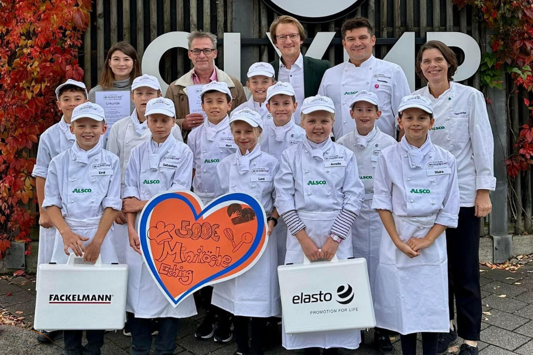 Eine Gruppe von Kindern in weißen Kochuniformen, begleitet von Erwachsenen, mit Spendenherz