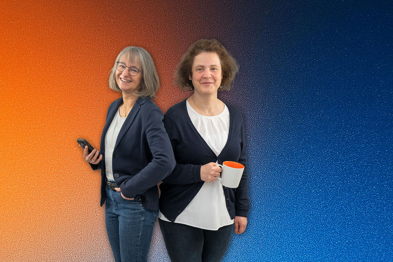 Zwei Barmstedter Kolleginnen stehen Rücken an Rücken, eine hält ein Smartphone , die andere eine Kaffeetasse