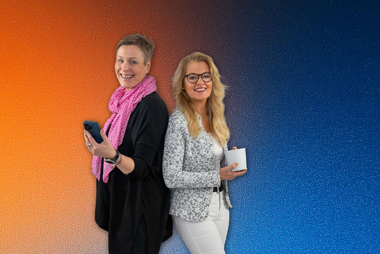 Zwei Ellerauer Kolleginnen stehen Rücken an Rücken, eine hält ein Smartphone , die andere eine Kaffeetasse