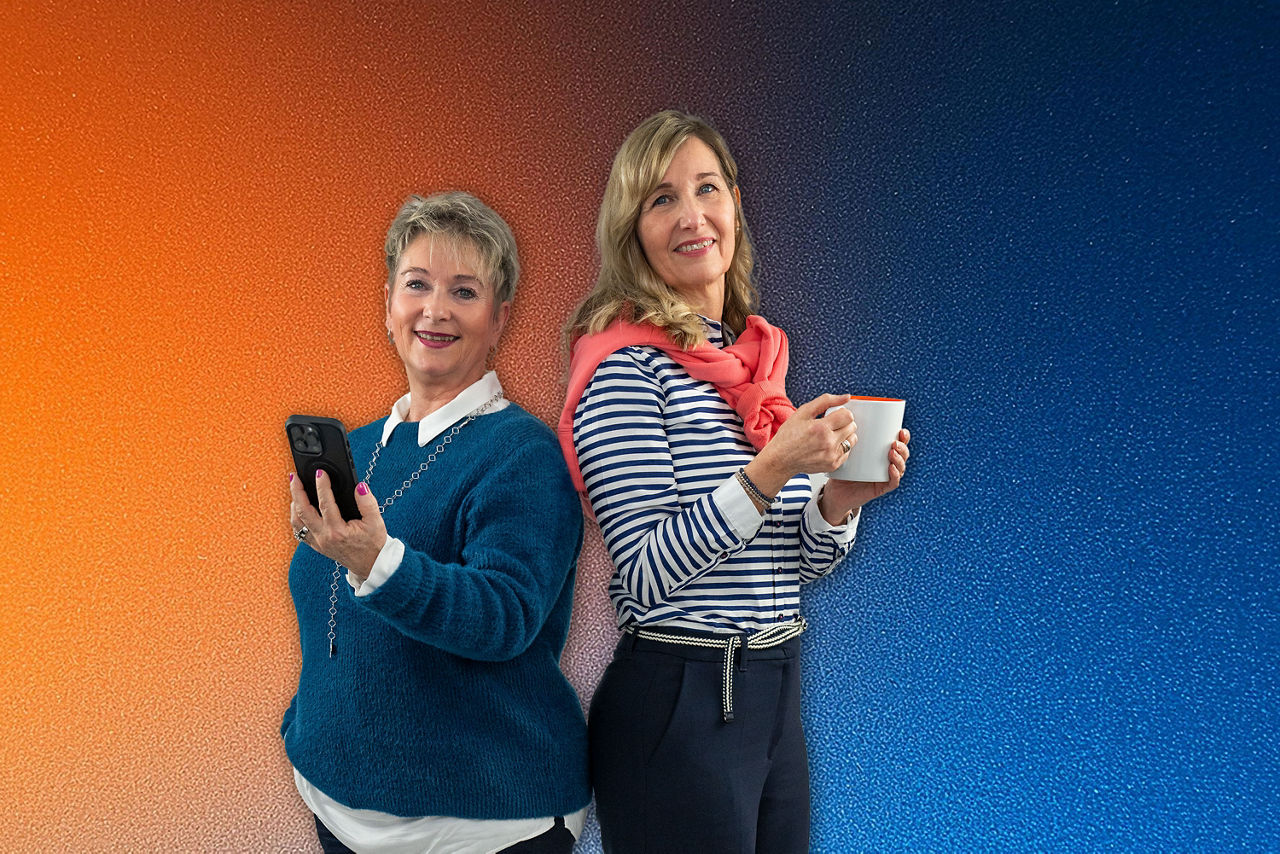 Zwei Quickborner Kolleginnen stehen Rücken an Rücken, eine hält ein Smartphone , die andere eine Kaffeetasse