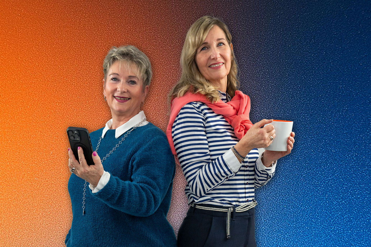 Zwei Quickborner Kolleginnen stehen Rücken an Rücken, eine hält ein Smartphone , die andere eine Kaffeetasse