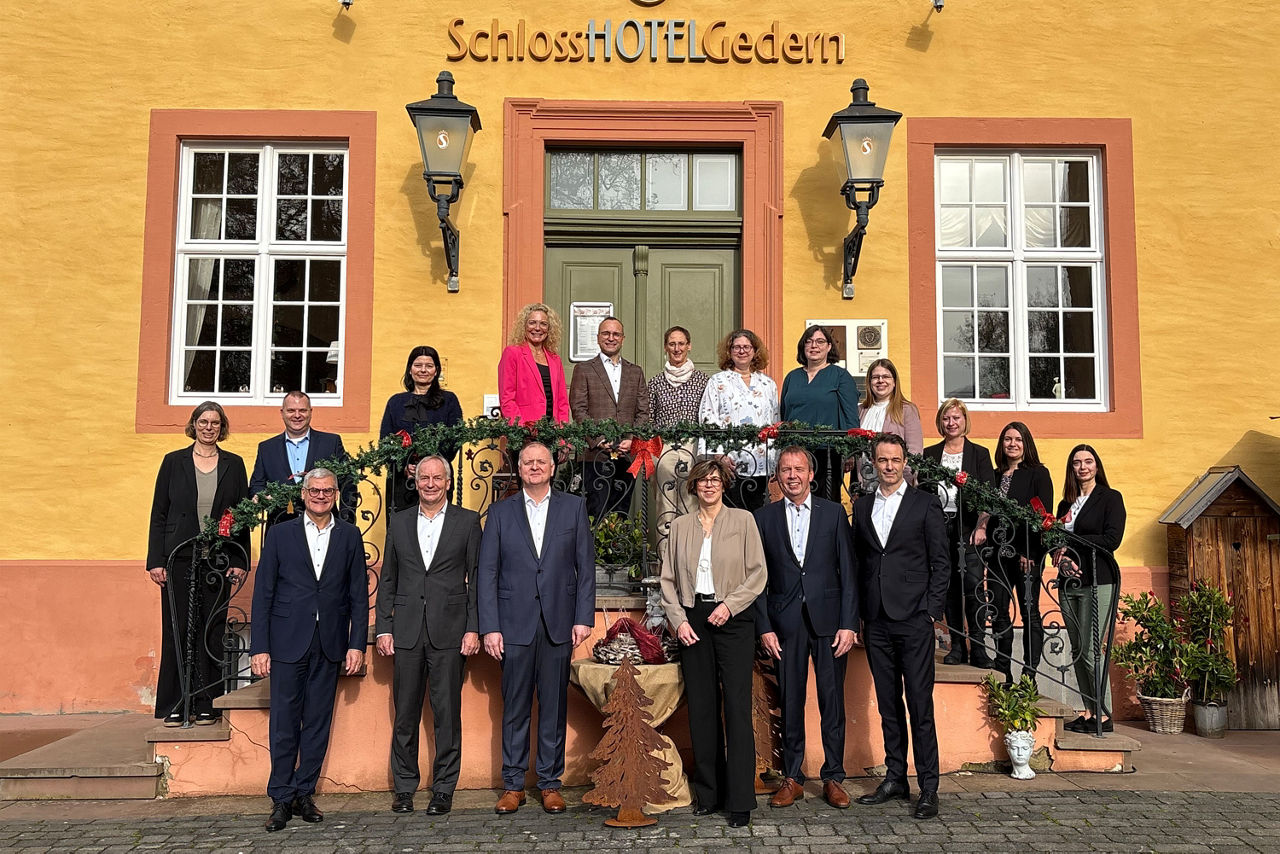 Eine Gruppe von 18 Personen steht vor einem gelben Gebäude mit der Aufschrift „SchlossHOTEL Gedern“. Einige stehen auf einer Treppe, andere davor. Die Personen tragen formelle Kleidung. Die Treppe ist weihnachtlich geschmückt.
