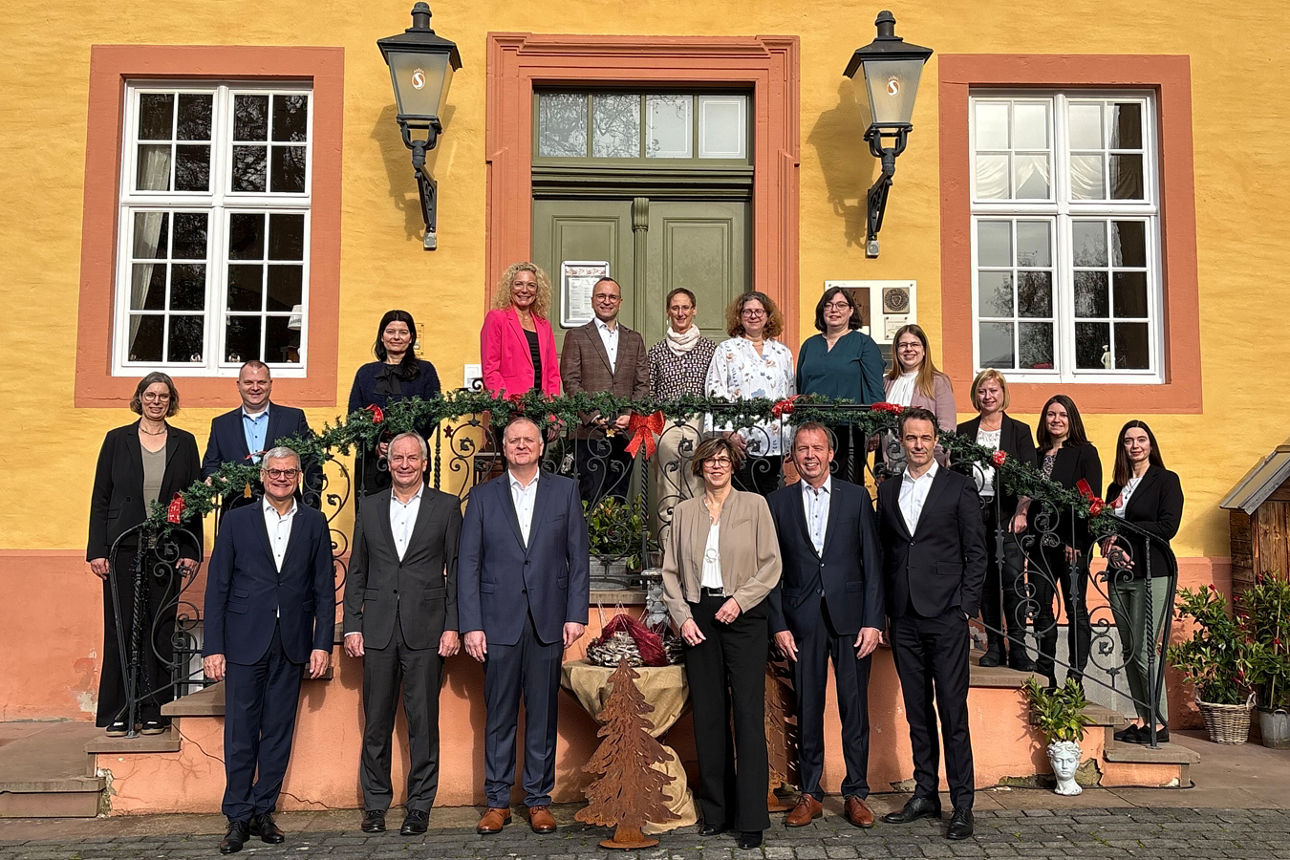 Eine Gruppe von 18 Personen steht vor einem gelben Gebäude mit der Aufschrift „SchlossHOTEL Gedern“. Einige stehen auf einer Treppe, andere davor. Die Personen tragen formelle Kleidung. Die Treppe ist weihnachtlich geschmückt.