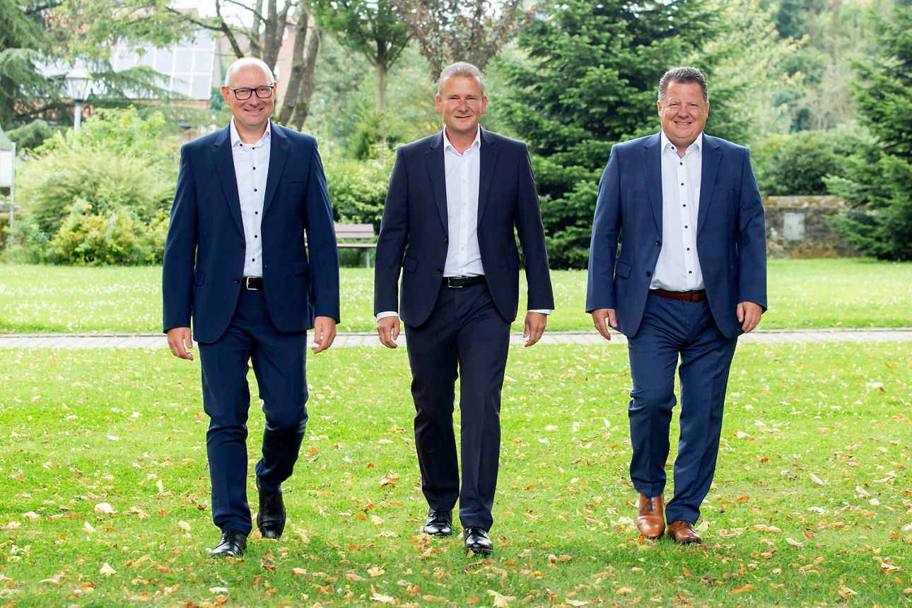 Drei ältere Männer im Anzug gehen auf einer Wiese auf die Kamera zu