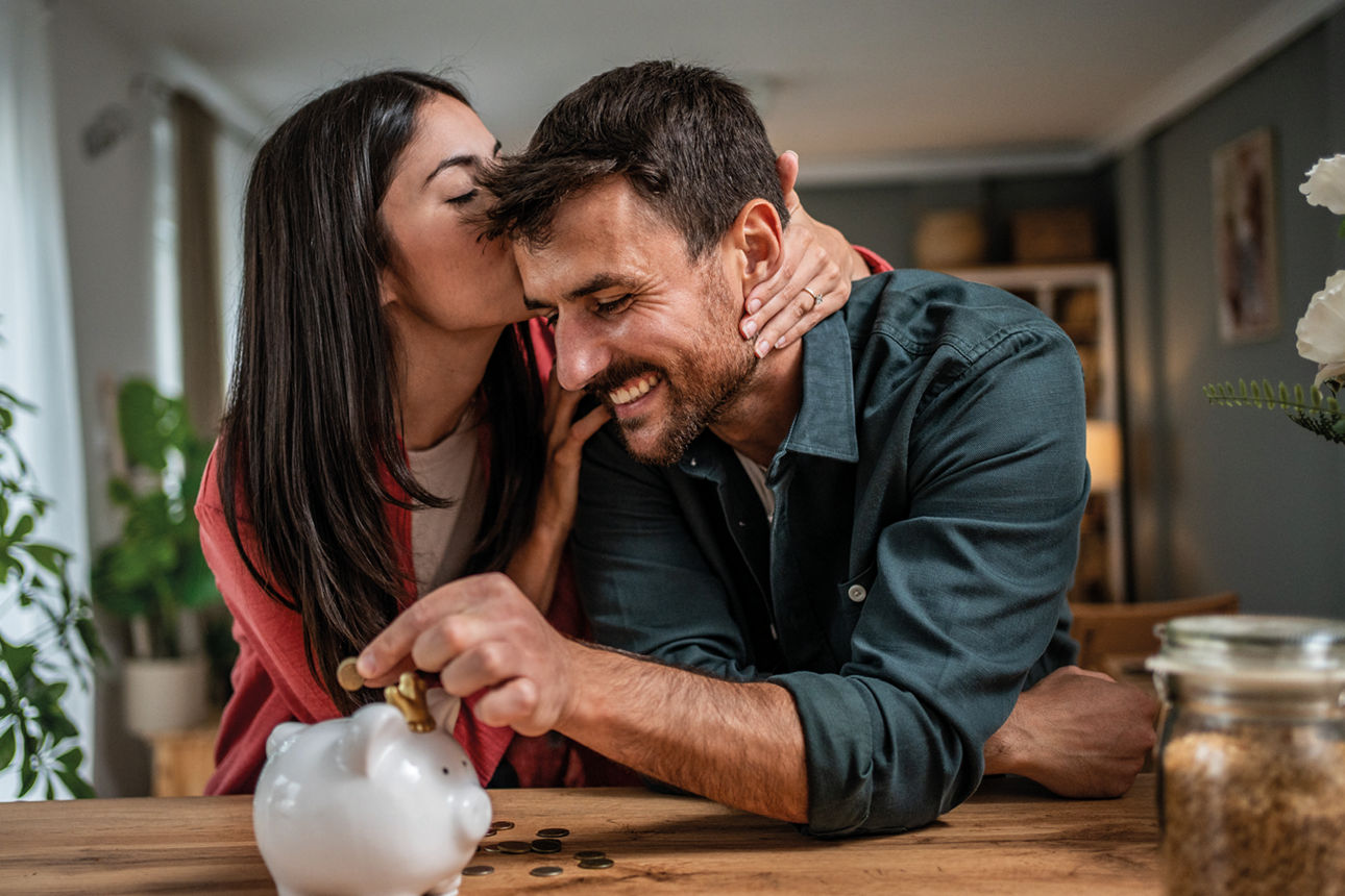 Pärchen am Küchentisch wo Sie Ihn an die Stirn küsst während er Münzen in ein Sparschwein steckt.