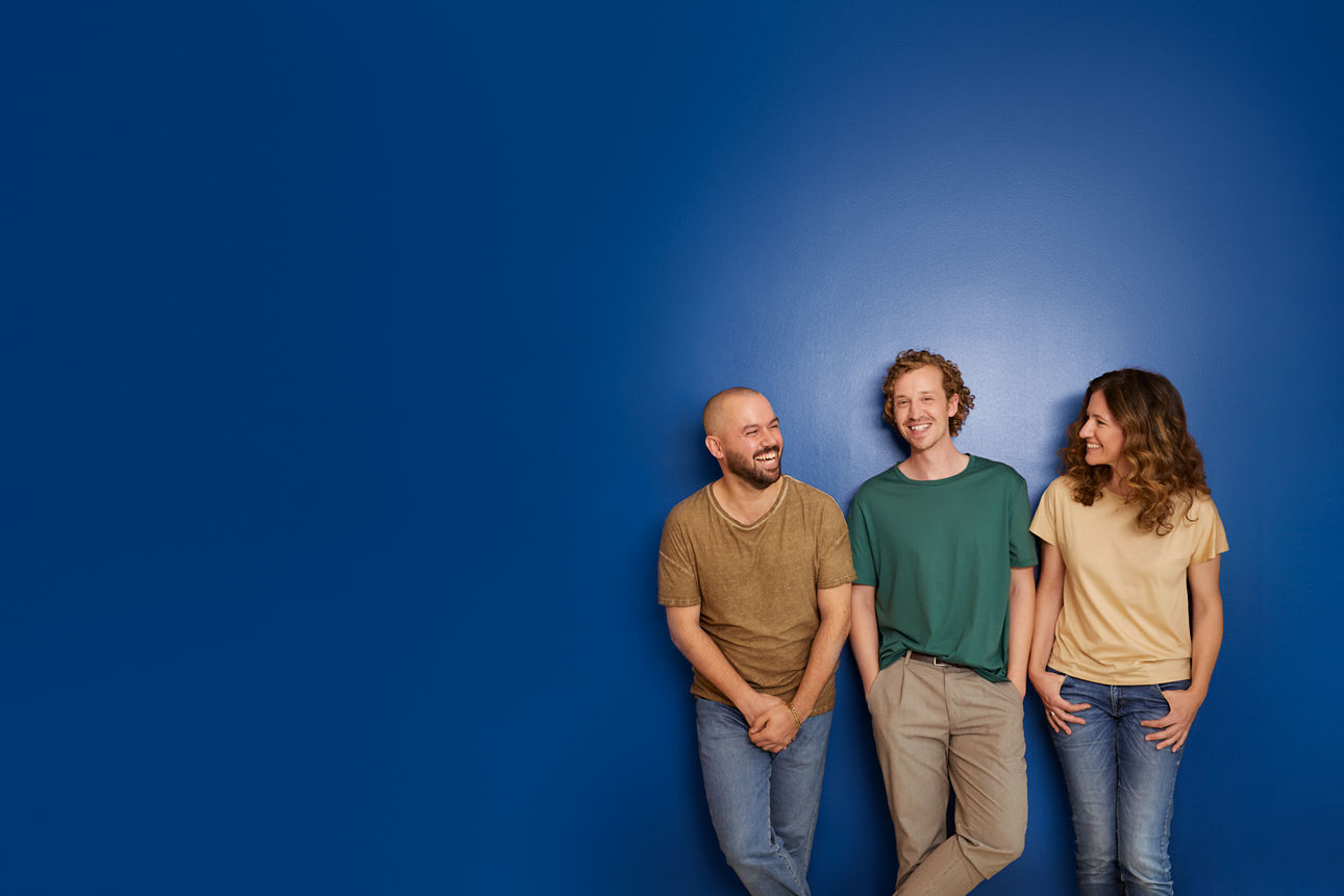 Drei Personen eine Frau und zwei Männer, angelehnt an einer blauen Wand. Alle Lächeln, die beiden Äußeren schauen sich an.