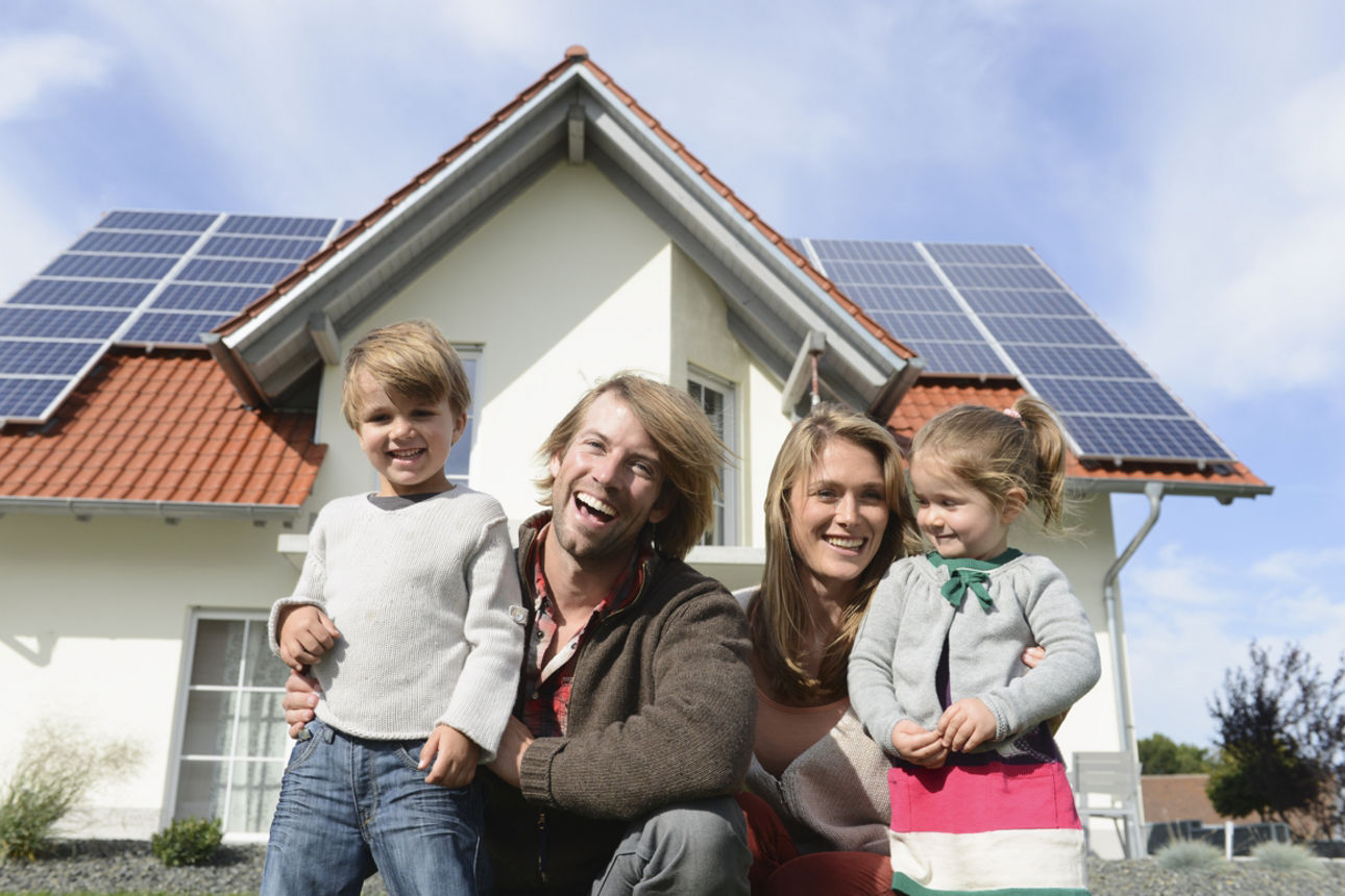 Eine Frau, ein Mann und zwei Kinder sind lachend vor einem Haus, welches eine PV-Anlage auf dem Dach hat