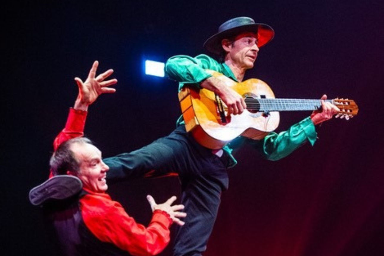 Auf dem Bild steht ein Künstler mit Gitarre auf dem Oberschenkel eines weiteren Künstlers