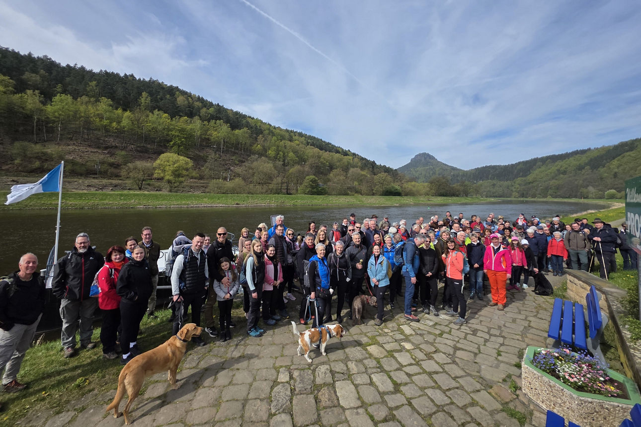 150 Personen in hügeliger Natur am Kanal in Wanderkleidung mit einigen Hunden