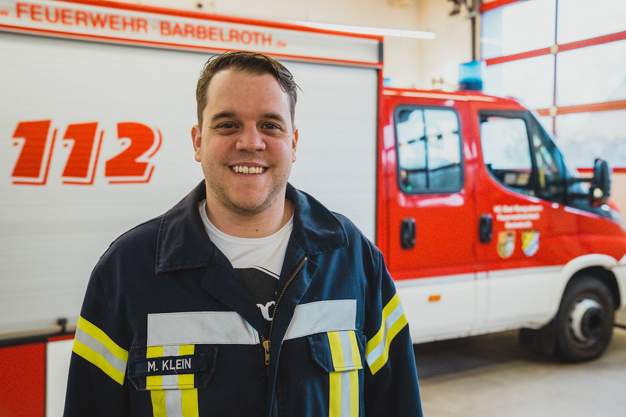 Feuerwehrmann steht lächelnd in Uniform vor einem Einsatzfahrzeug der Freiwilligen Feuerwehr in der Fahrzeughalle.