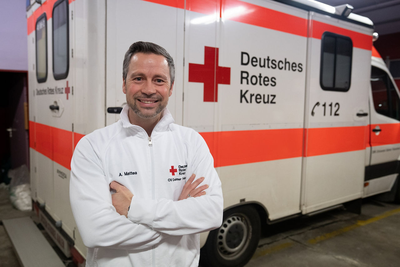 Mitarbeiter des Deutschen Roten Kreuzes steht lächelnd vor einem Einsatzfahrzeug mit DRK-Logo in einer Garage.